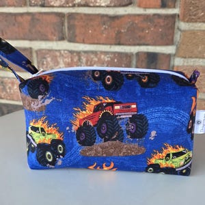 May include: A blue zippered pouch with a repeating pattern of monster trucks. The trucks are red, green, and black, with flames and large tires. The pouch has a small loop handle and a white zipper.