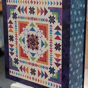 May include: A rectangular storage box featuring a vibrant quilt pattern on the front with multi-colored geometric shapes, stars, and triangles. The side panel is dark blue with light blue polka dots. The box displays the Anna Maria Textiles logo.