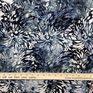 May include: Fabric with a blue and white floral pattern. The design features stylized, abstract flowers and foliage in shades of blue, gray, and white. A wooden ruler with text is at the bottom.
