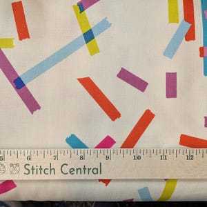 May include: White fabric with a colorful abstract design. The pattern features overlapping brushstrokes in shades of red, orange, yellow, blue, and purple. A wooden ruler with the text "Stitch Central" is at the bottom.