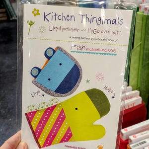 May include: A sewing pattern for a Lloyd potholder and Hugo oven mitt, featuring whimsical designs. The pattern includes finished sizes: Lloyd 11.5" x 6.5" and Hugo 12.5" x 8". The package is labeled "Kitchen Thingimals" and "Fish museum+circus".