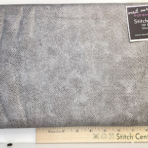 May include: Grey fabric featuring a repeating hexagonal design. A ruler with inch markings is at the bottom. A tag in the upper right corner reads "visit our new forever home! Stitch Central".