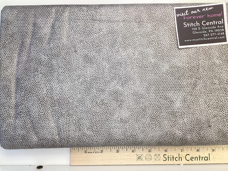 May include: Grey fabric featuring a repeating hexagonal design. A ruler with inch markings is at the bottom. A tag in the upper right corner reads "visit our new forever home! Stitch Central".