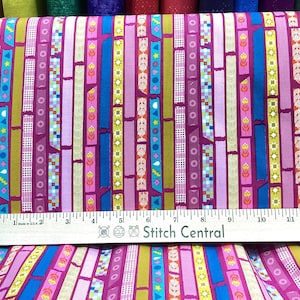 May include: A roll of fabric with a repeating vertical pattern of colorful ribbons and rectangles. The ribbons are in shades of pink, blue, green, and yellow, with floral and geometric designs. A wooden ruler is placed on the fabric.