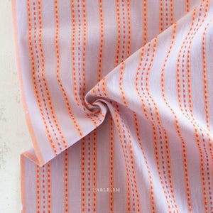 May include: A close-up of a fabric with vertical orange dotted lines on a light purple background. The fabric is folded, showing the texture and pattern. The word "FABLEISM" is printed in the bottom right corner.