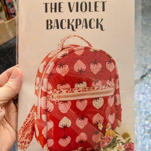 May include: A red backpack pattern with a strawberry design, titled "THE VIOLET BACKPACK." The backpack has a zippered front pocket and a floral print on the straps and zipper. The pattern is in a clear plastic sleeve.