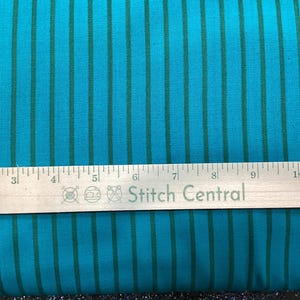 May include: A vibrant turquoise fabric featuring thin, vertical dark green stripes. A metal ruler is positioned across the fabric, showing measurements in inches with the text Stitch Central printed on the surface of the ruler.