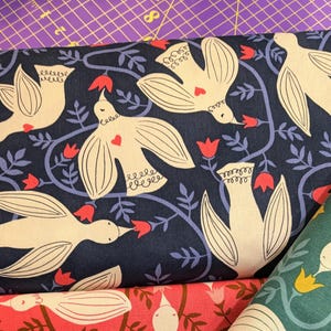 May include: A navy blue fabric with a repeating pattern of white birds and pink flowers. The birds are stylized and have a heart shape on their chest. The fabric is perfect for quilting, sewing, and crafting.