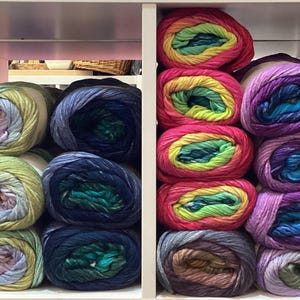 May include: Two shelves filled with colorful yarn skeins. The top shelf has rainbow colored yarn skeins and the bottom shelf has yarn skeins in shades of blue, green, and purple.