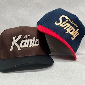 Może przedstawiać: Dwie czapki bejsbolowe. Jedna jest brązowa z czarnym daszkiem i napisana "VISIT KANTO" białym kursywą. Druga jest granatowa z czerwonym daszkiem i napisana "Simply COLLECTABLE" białym kursywą ze złotymi akcentami.