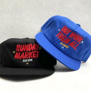 Może przedstawiać: Dwie czapki bejsbolowe, jedna czarna i jedna niebieska. Czarna czapka ma czerwony haft z napisem "SUNDAY MARKET ROSE BOWL". Niebieska czapka ma czerwony haft z napisem "OUT WORK THEM ALL LOS ANGELES".