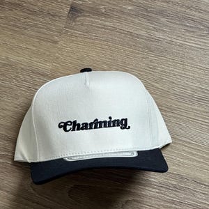 May include: A white and black baseball cap with the word "Charming" embroidered in black cursive on the front.