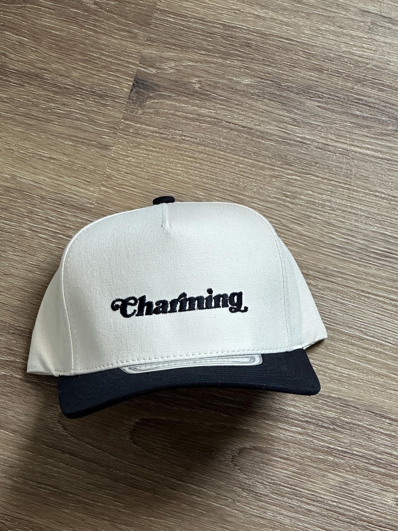 May include: A white and black baseball cap with the word "Charming" embroidered in black cursive on the front.