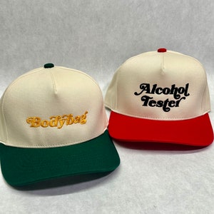 May include: Two baseball caps with different coloured brims. One cap is white with a green brim and says "Bodybag" in gold embroidery. The other cap is white with a red brim and says "Alcohol Tester" in black embroidery.