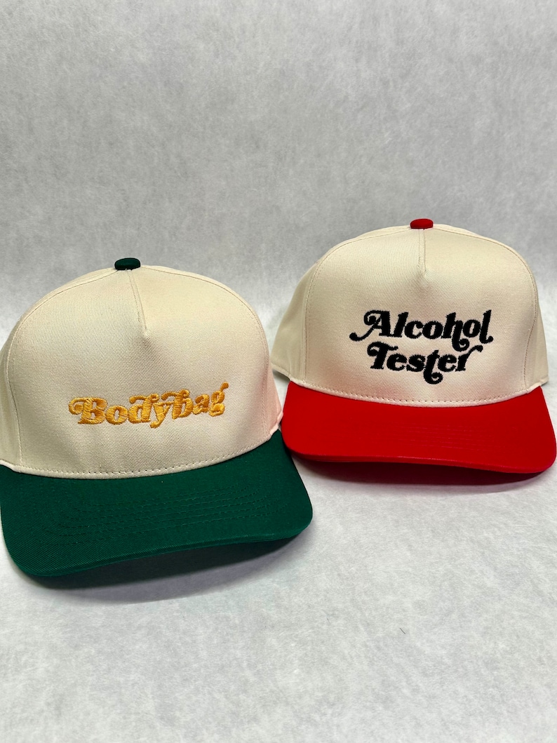May include: Two baseball caps with different coloured brims. One cap is white with a green brim and says "Bodybag" in gold embroidery. The other cap is white with a red brim and says "Alcohol Tester" in black embroidery.