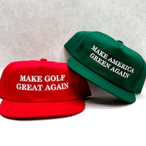 Könnte beinhalten: Zwei Baseballkappen. Eine ist rot mit dem weißen Text "MAKE GOLF GREAT AGAIN". Die andere ist grün mit dem weißen Text "MAKE AMERICA GREEN AGAIN". Beide Kappen haben einen flachen Schirm und einen Knopf oben.