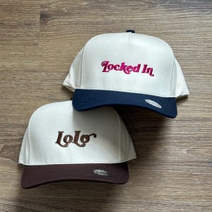 May include: Two white baseball caps with different coloured brims and embroidered text. The top cap has a navy blue brim and says "Locked In" in pink. The bottom cap has a brown brim and says "lolo" in brown.