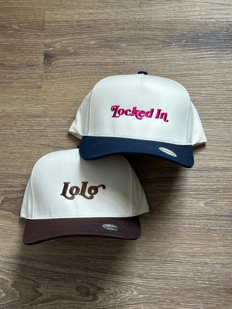 May include: Two white baseball caps with different coloured brims and embroidered text. The top cap has a navy blue brim and says "Locked In" in pink. The bottom cap has a brown brim and says "lolo" in brown.