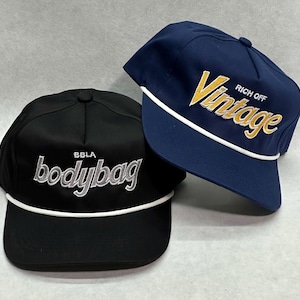 May include: Two baseball caps. One is black with "bodybag" embroidered in silver, and the other is navy blue with "Vintage" in yellow. Both have a white rope detail along the brim.