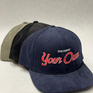 May include: Three corduroy baseball caps in olive green, black, and navy blue. The navy cap features the text "Customize Your Own" in red script. Each cap has a curved brim and a top button.