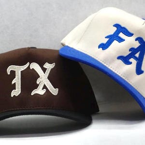以下が含まれることがあります： 2つの野球帽。1つは茶色で白い「TX」の文字、もう1つは白で青い「FA」の文字が入っています。