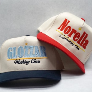 Puede incluir: Dos gorras de béisbol, una blanca con ribete rojo y el texto "Norella Smoking Club" bordado en rojo y amarillo, y la otra blanca con ribete azul marino y el texto "Glozzar Working Class" bordado en azul y amarillo.