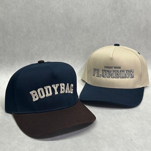 May include: Two baseball caps with embroidered text. One cap is navy blue with a brown brim and says "BODYBAG" in white. The other cap is beige with a navy blue brim and says "PLUMBING" in white with "VINCENT WONG" above it.