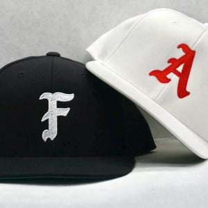 Puede incluir: Dos gorras de béisbol, una negra con una "F" blanca y una blanca con una "A" roja.