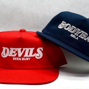 May include: Two baseball caps, one red with white embroidery that says "DEVILS BEEN BUSY" and one navy blue with white embroidery that says "BODYBAG BBLA".