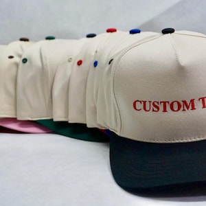 Puede incluir: Una colección de gorras de béisbol beige con viseras negras, cada una con un botón de diferente color en la parte superior. La gorra delantera tiene "CUSTOM TEXT" bordado en rojo.