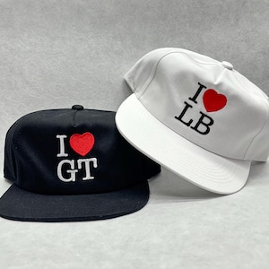 以下が含まれることがあります： フラットブリムの野球帽が2つ。1つは黒で、白と赤で「I ❤️ GT」と刺繍されています。もう1つは白で、赤と黒で「I ❤️ LB」と刺繍されています。どちらの帽子にも上部にボタンが付いています。