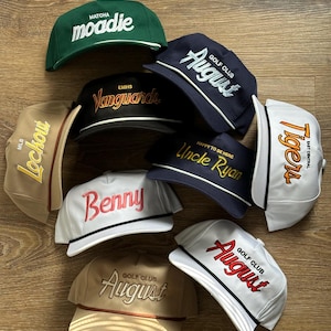 May include: A selection of baseball caps in shades like green, navy, beige, and white. Each cap displays embroidered text, including "moadie," "August," "Benny," and "Tigers." The caps are arranged on a wooden surface.