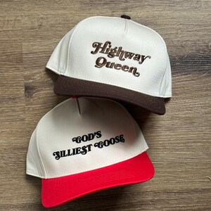 May include: Two white baseball caps with brown and red brims. The front of the caps have embroidered text. The top cap says "Highway Queen" in brown cursive text. The bottom cap says "God's Silliest Goose" in black cursive text.