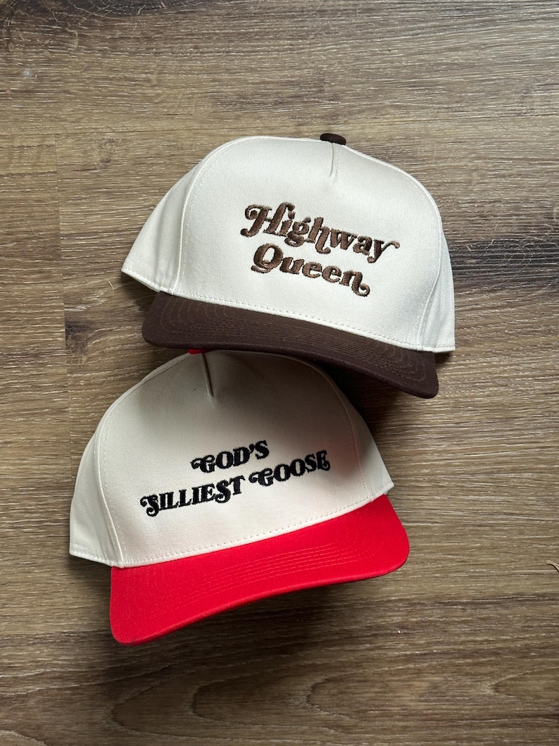 May include: Two white baseball caps with brown and red brims. The front of the caps have embroidered text. The top cap says "Highway Queen" in brown cursive text. The bottom cap says "God's Silliest Goose" in black cursive text.