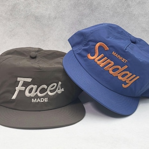 May include: A dark gray baseball cap with white embroidered text reading Faces MADE and a blue baseball cap with orange embroidered text reading Sunday MARKET. Both caps feature a structured crown, a button top, and a flat brim design.