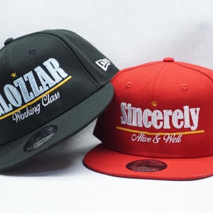 May include: Two baseball caps, one black and one red. The black cap has the word "GLOZZAR" in white, with "Working Class" below. The red cap says "Sincerely Alive & Well" in white. Both have a New Era logo.