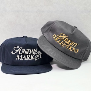 以下が含まれることがあります： SUNDAY MARKETと白で刺繍されたネイビーの野球帽と、BRIGHT COLLECTIONSとゴールドで刺繍されたチャコールグレーの野球帽。どちらの帽子も構造化されたクラウンとフラットなつばのデザインが特徴です。