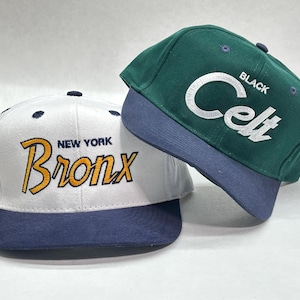 May include: Two baseball caps. One is white with "NEW YORK Bronx" in yellow and navy blue. The other is green with "BLACK Celt" in white. Both have navy blue brims.