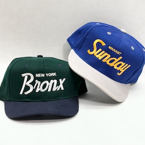 May include: Two baseball caps. One is dark green with "NEW YORK Bronx" in white lettering. The other is blue with "MARKET Sunday" in yellow lettering and a white brim. Both caps have a flat brim.