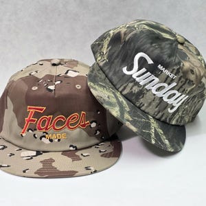 May include: Two baseball caps with flat brims. The left cap features a tan and brown camouflage pattern with the red embroidered text "Faces" and yellow "MADE" below it. The right cap has a green and black forest camouflage pattern with white "Sunday MARKET" text.