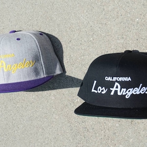 Pode incluir: Duas bonés de beisebol com aba plana e coroa curva. O boné à esquerda é cinza com aba roxa e o texto "CALIFORNIA Los Angeles" em bordado amarelo. O boné à direita é preto com aba preta e o texto "CALIFORNIA Los Angeles" em bordado branco.