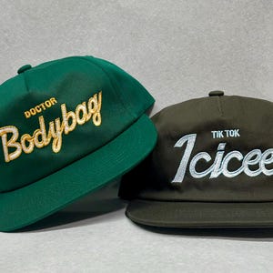 May include: Two baseball caps, one green with gold embroidery that says "Doctor Bodybag" and one brown with white embroidery that says "Tik Tok Icicee".