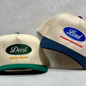 May include: Two beige baseball caps with embroidered text. One cap has a green brim and the word "Devil" in white script within a green oval, with "BEEN BUSY" below in gold. The other cap has a navy brim and the word "Lord" in blue within a blue oval, with "PLEASE FORGIVE ME" below in red.