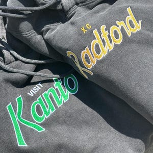 May include: Black hooded sweatshirt with green and white embroidery that says "VISIT KANTO" and yellow embroidery that says "Radford" with the letters "XC" above it.