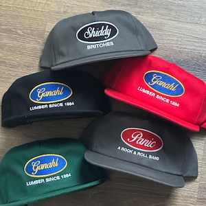 Może przedstawiać: Pięć czapek bejsbolowych w różnych kolorach, w tym szarym, czarnym, czerwonym i zielonym. Każda czapka ma haftowany tekst, z frazami takimi jak "Shiddy Britches", "Ganahl Lumber Since 1884" i "Panic A Rock & Roll Band".