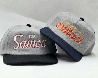 May include: Two grey baseball caps with flat brims. The left cap features white embroidered text reading TOA Samoa. The right cap features orange and red embroidered text reading artifact and CRAFTED BY LONG. Both caps have a small black button on top.