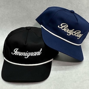 May include: Two baseball caps, one black and one navy blue. The black cap has the word "Immigrant" embroidered in white script. The navy blue cap has the words "Body Bag" embroidered in white script. Both caps have a white rope detail along the brim.
