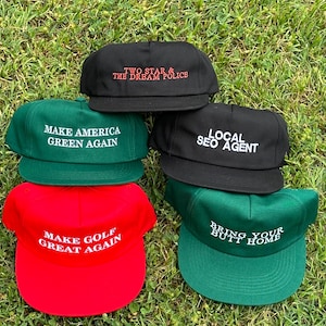 Op de afbeelding: Een verzameling baseballpetten in verschillende kleuren, waaronder zwart, rood en groen. Elke pet heeft witte tekst met zinnen als "Make America Green Again" en "Make Golf Great Again". De petten zijn gestapeld op groen gras.