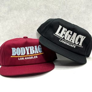 Puede incluir: Dos gorras de béisbol. Una es negra con "LEGACY LANDSCAPE" bordado en blanco. La otra es burdeos con "BODYBAG LOS ANGELES" bordado en azul claro y amarillo. Ambas gorras tienen visera plana y un ajuste estructurado.