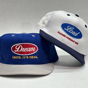 Puede incluir: Dos gorras de béisbol. Una es azul con la palabra "Dream" en un óvalo rojo y el texto "UNTIL IT'S REAL". La otra es blanca con un óvalo azul que dice "Lord" y el texto "PLEASE FORGIVE ME".